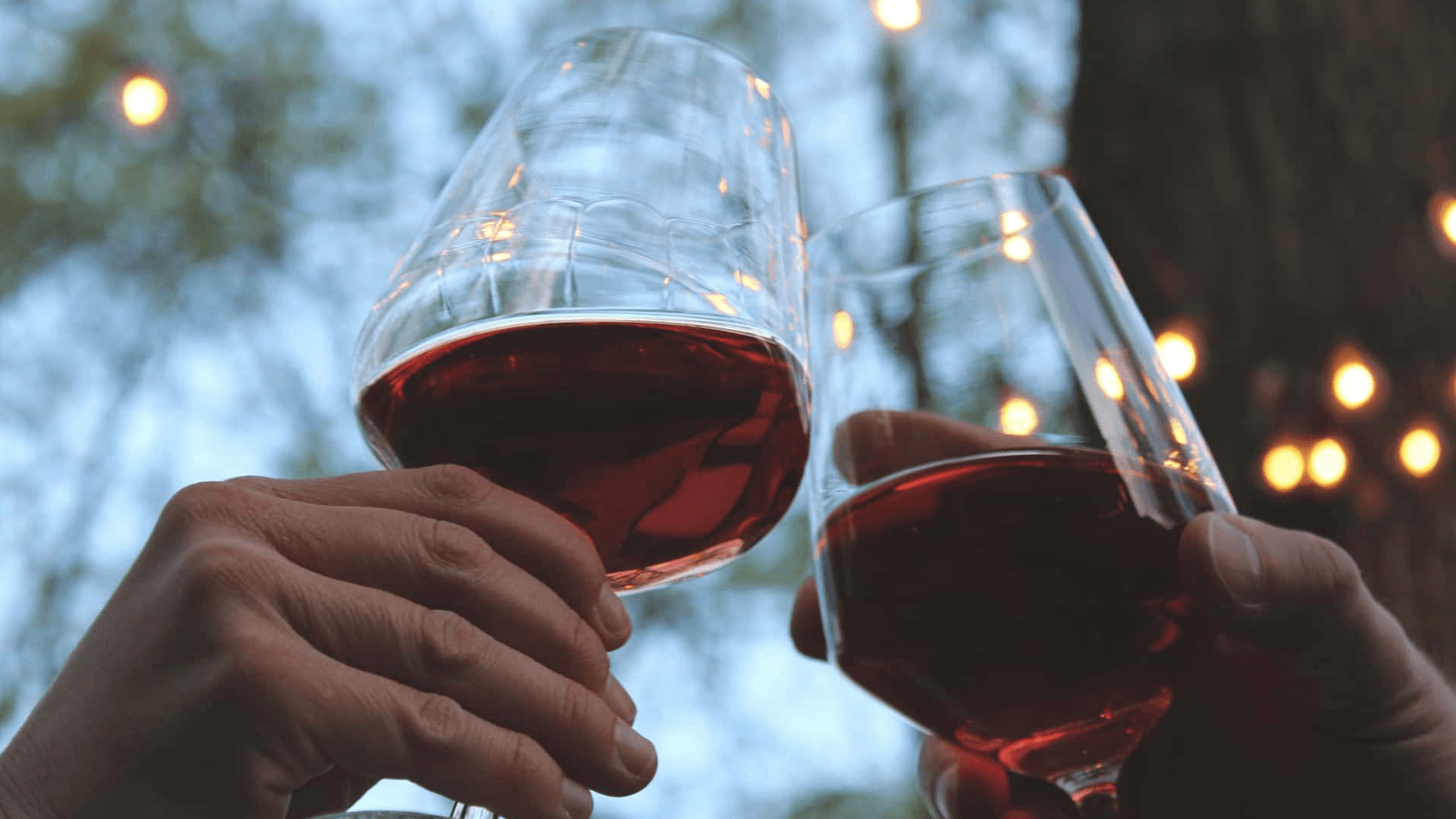 Two hands clinking wine glasses filled with red wine against a softly lit outdoor background.