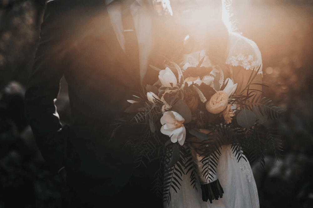 A bride and groom embrace, with the bride holding a lush bouquet in soft sunlight.