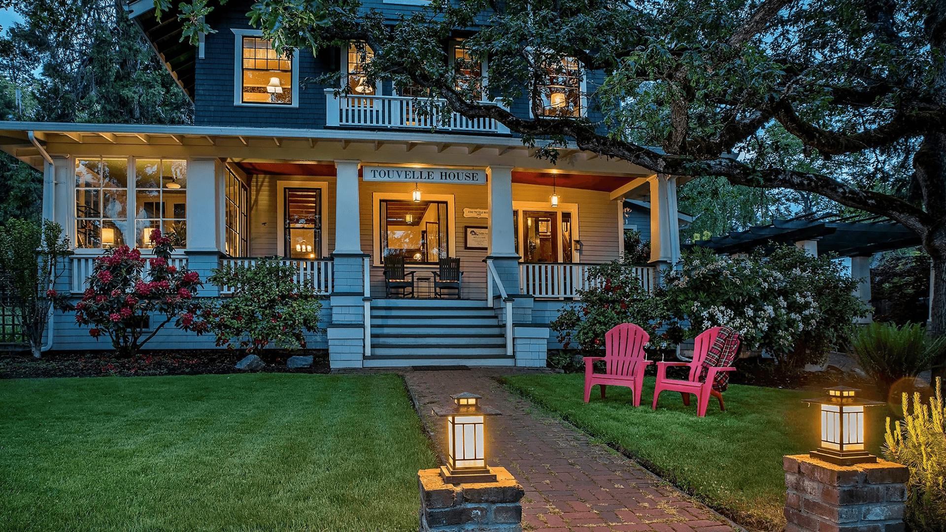 A welcoming two-story house with a porch, surrounded by greenery and illuminated by lamps, features two red chairs on the lawn.